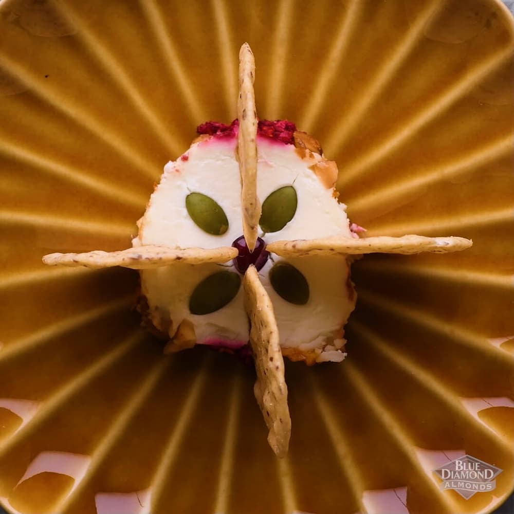 A round slice of cheese topped with four pumpkin seeds and surrounded by four almond crackers, arranged on a brown, radiating patterned plate.