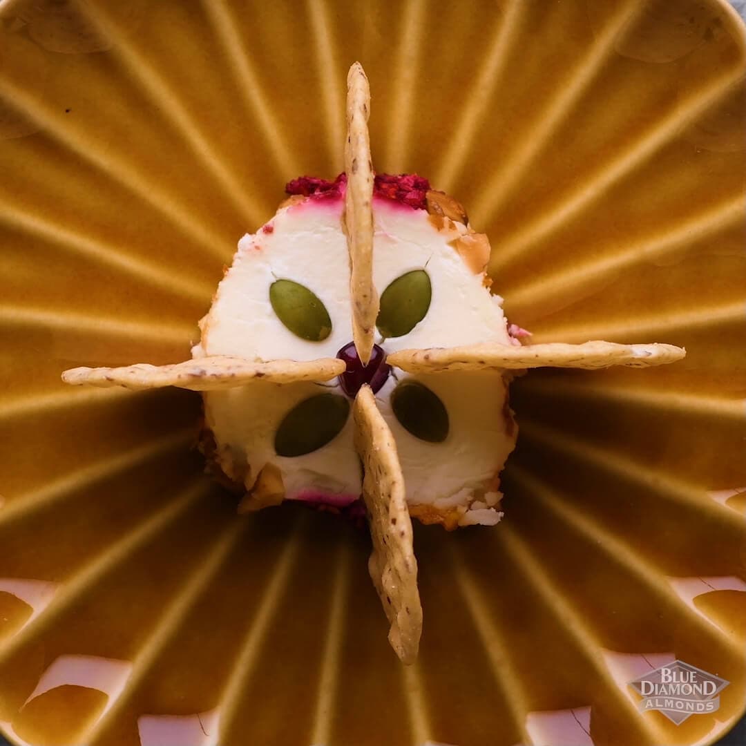 A round slice of cheese topped with four pumpkin seeds and surrounded by four almond crackers, arranged on a brown, radiating patterned plate.