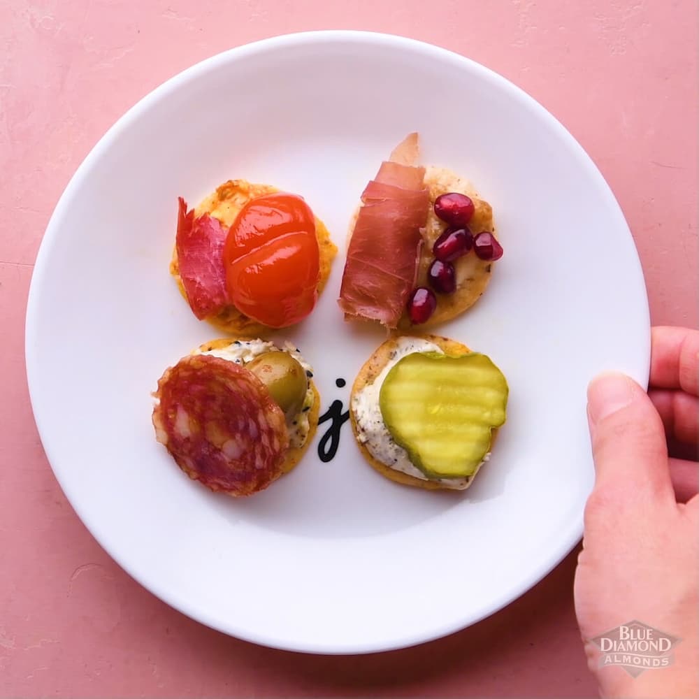 A white plate with four assorted crackers topped with meats, cheese, tomato, pomegranate seeds, and pickles, on a pink background. A hand is holding the edge of the plate.