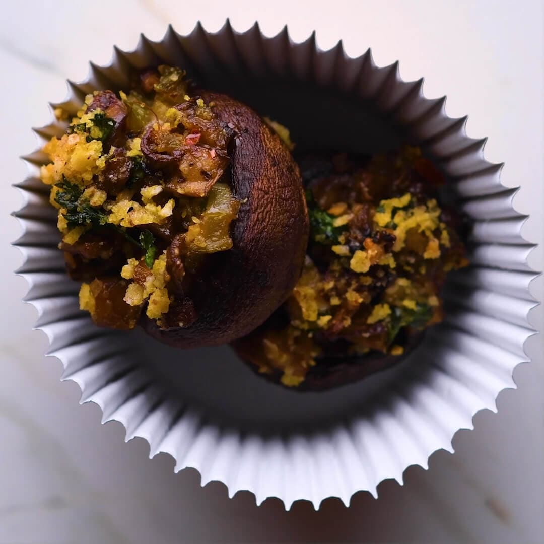 Two stuffed mushrooms with a crumb topping sit inside a white paper baking cup, viewed from above on a light-colored surface.