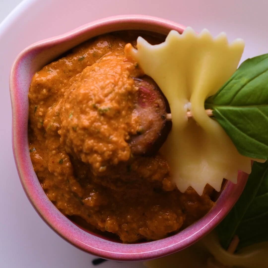 A small bowl of thick red sauce with a slice of sausage, garnished with a piece of farfalle pasta and a fresh basil leaf.