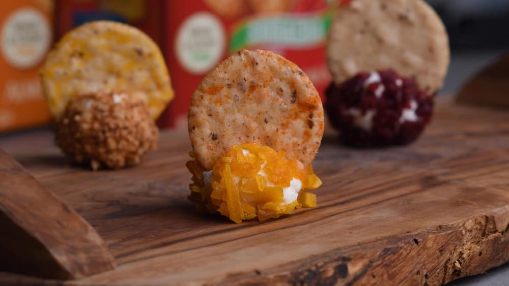Round crackers topped with various cheese spreads and coatings are arranged on a wooden board with blurred product boxes in the background.