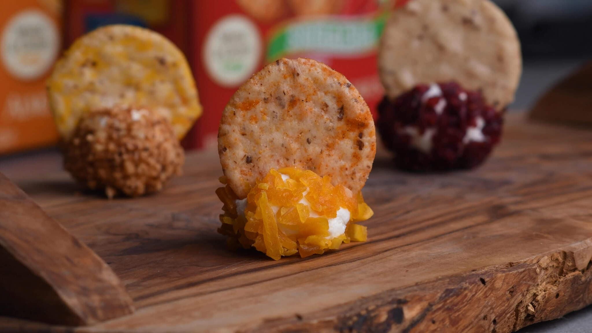 Round crackers topped with various cheese spreads and coatings are arranged on a wooden board with blurred product boxes in the background.
