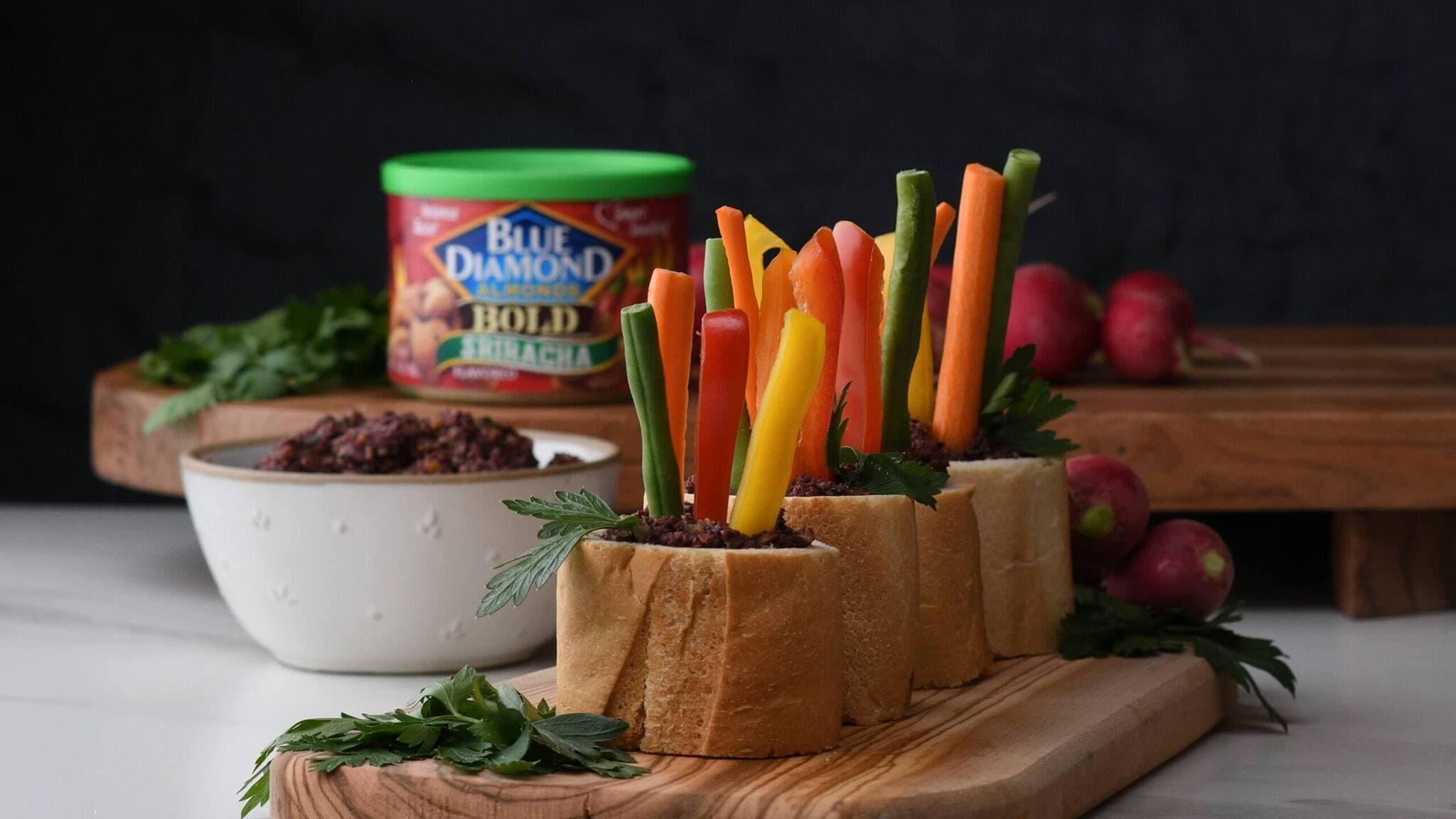 Vegetable sticks in bread rolls filled with dip are arranged on a wooden board. A bowl of dip and a can of Blue Diamond almonds sit in the background, along with fresh parsley and radishes.