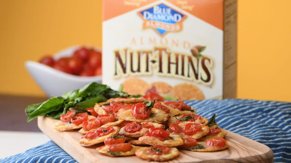 A wooden board with crackers topped with tomatoes and herbs, a bunch of leafy greens, and a Nut-Thins almond cracker box in the background.