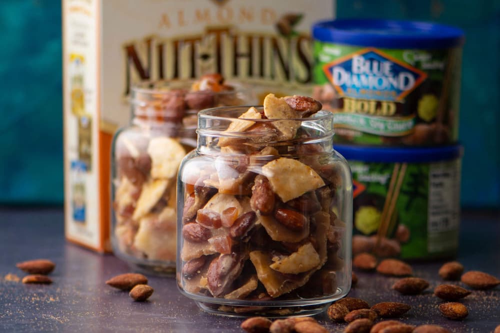 Two glass jars filled with nut and cracker snack mix are in front of Nut-Thins and Blue Diamond Almonds containers, with scattered almonds on the dark surface.