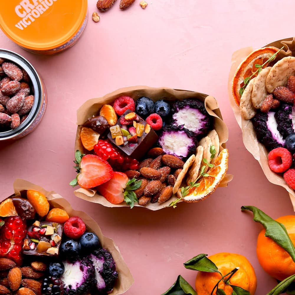 Three paper trays filled with assorted fruits, nuts, crackers, chocolates, and cheese are arranged on a pink surface, alongside a jar of almonds and whole mandarins with leaves.