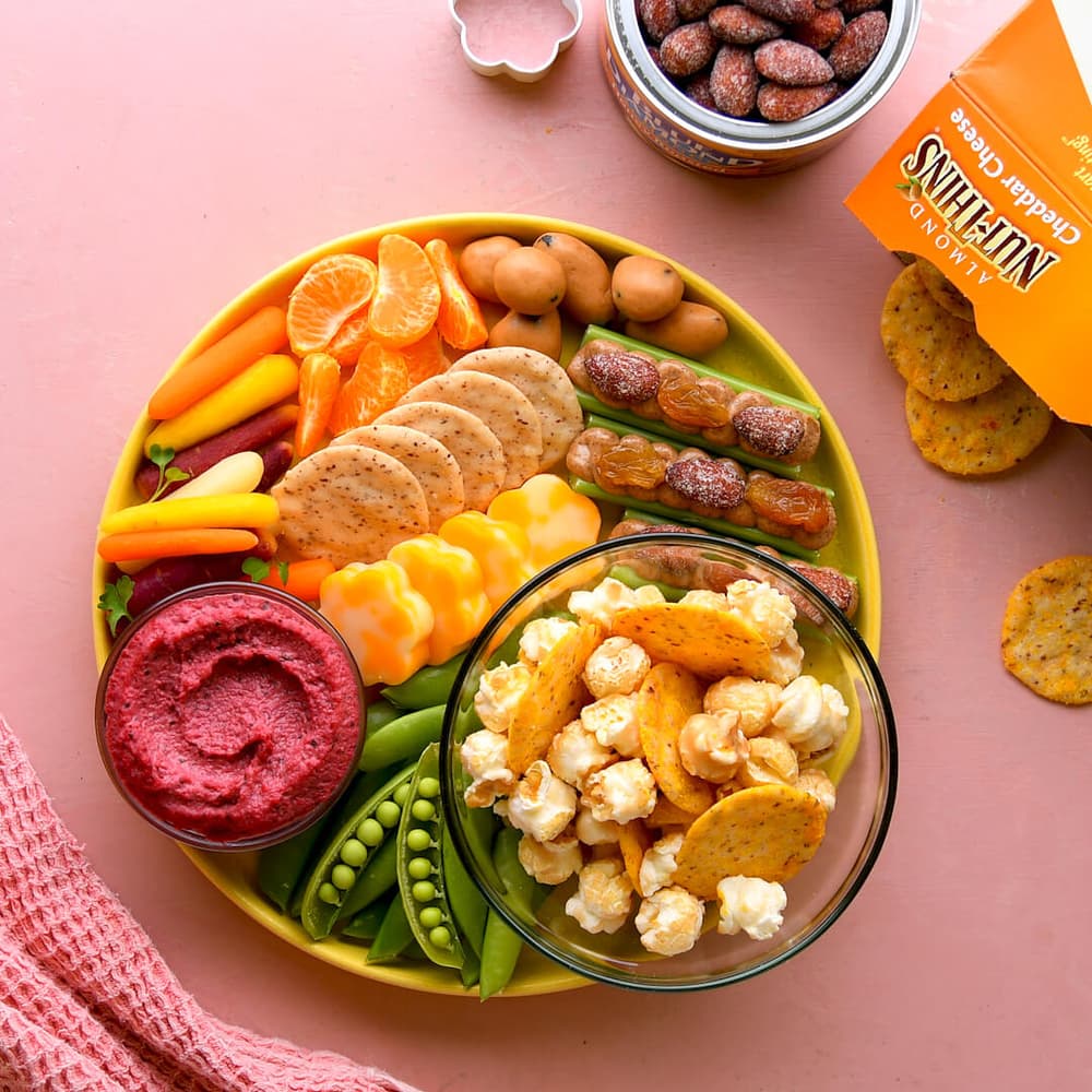 A yellow platter with crackers, cheese, mini sausages, carrot sticks, sugar snap peas, tangerine slices, beet dip, and a bowl of popcorn with cheese crisps. Almond container nearby.