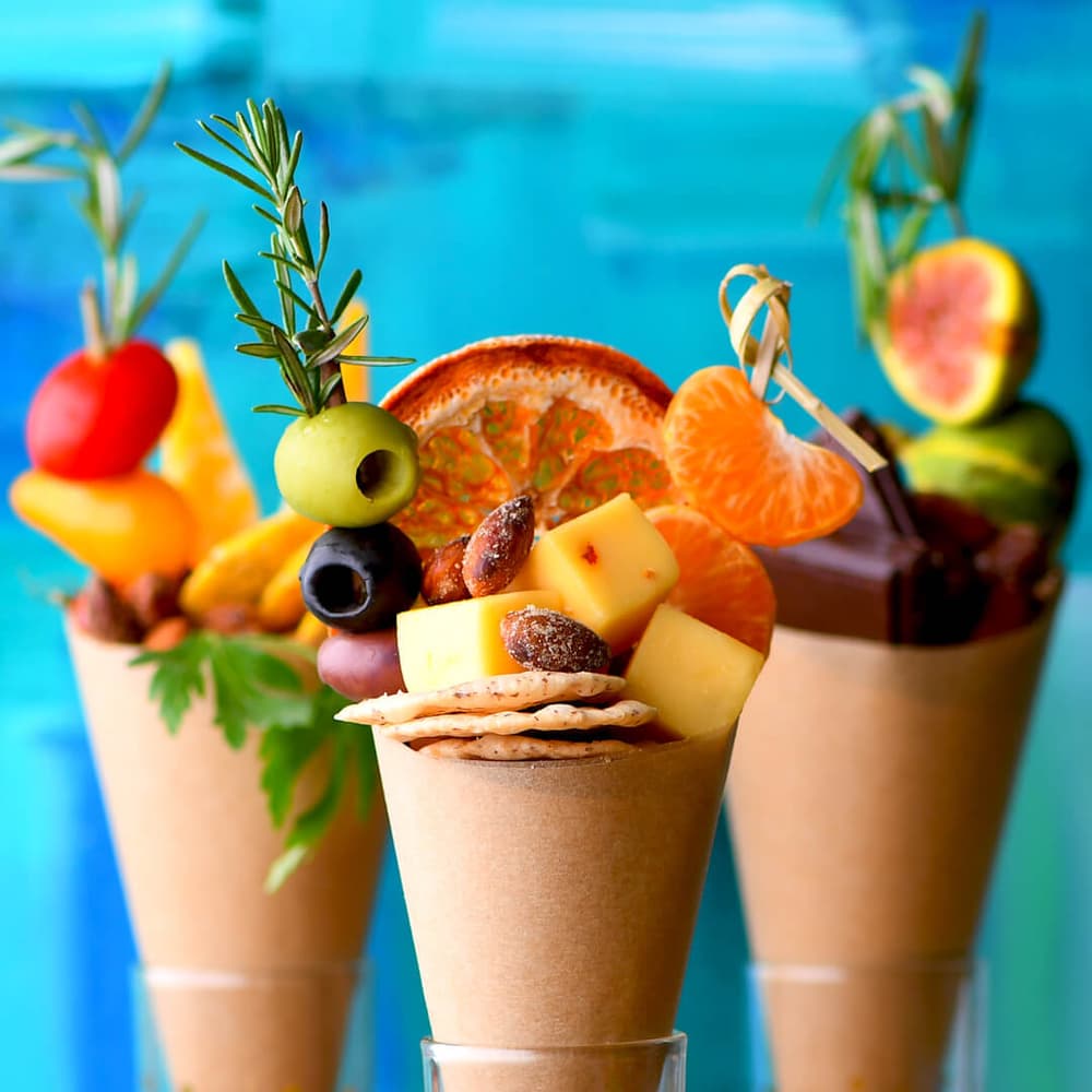 Three paper cones filled with assorted cheese cubes, crackers, fruit, olives, nuts, and herbs against a vibrant blue background.
