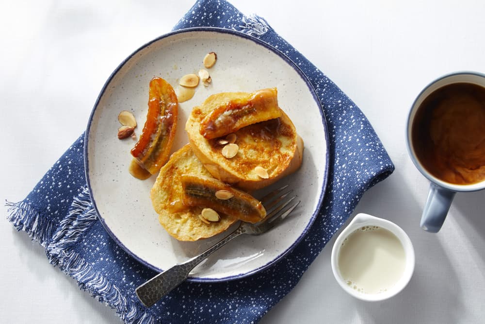 A plate with French toast topped with caramelized bananas and almonds, a fork, a cup of coffee, a small pitcher of milk, and a blue napkin on a white surface.