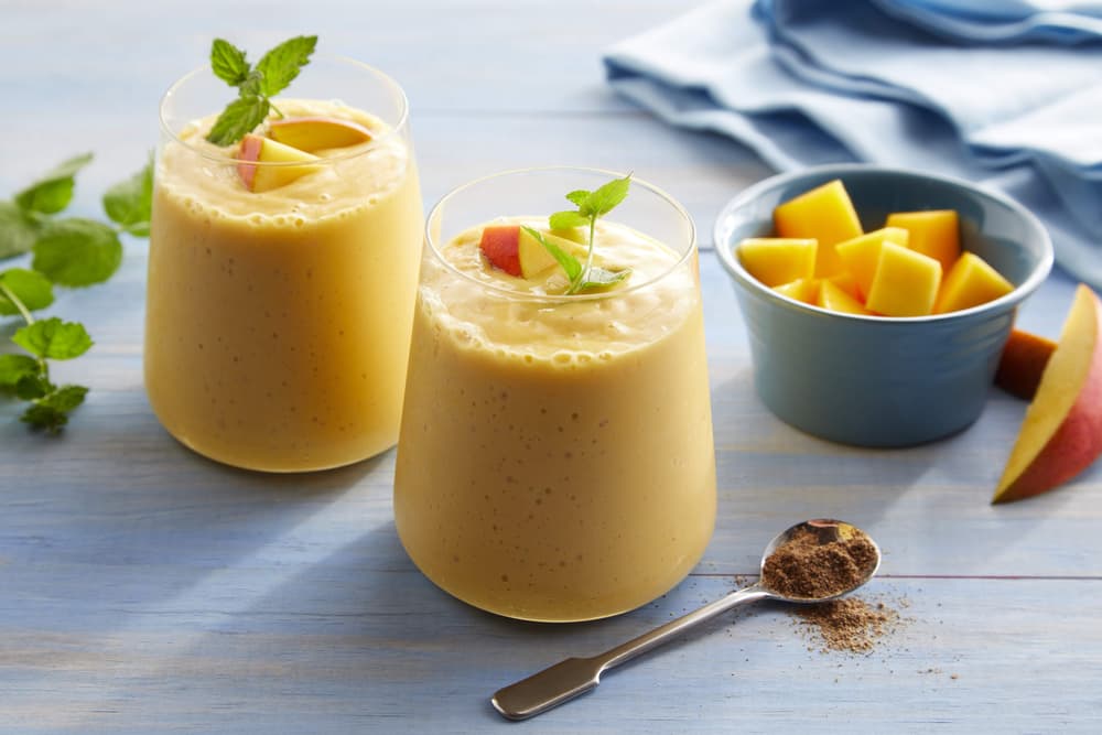 Two glasses of mango smoothie topped with mint and mango slices, next to a bowl of diced mango and a spoon with ground spice, on a light blue table.