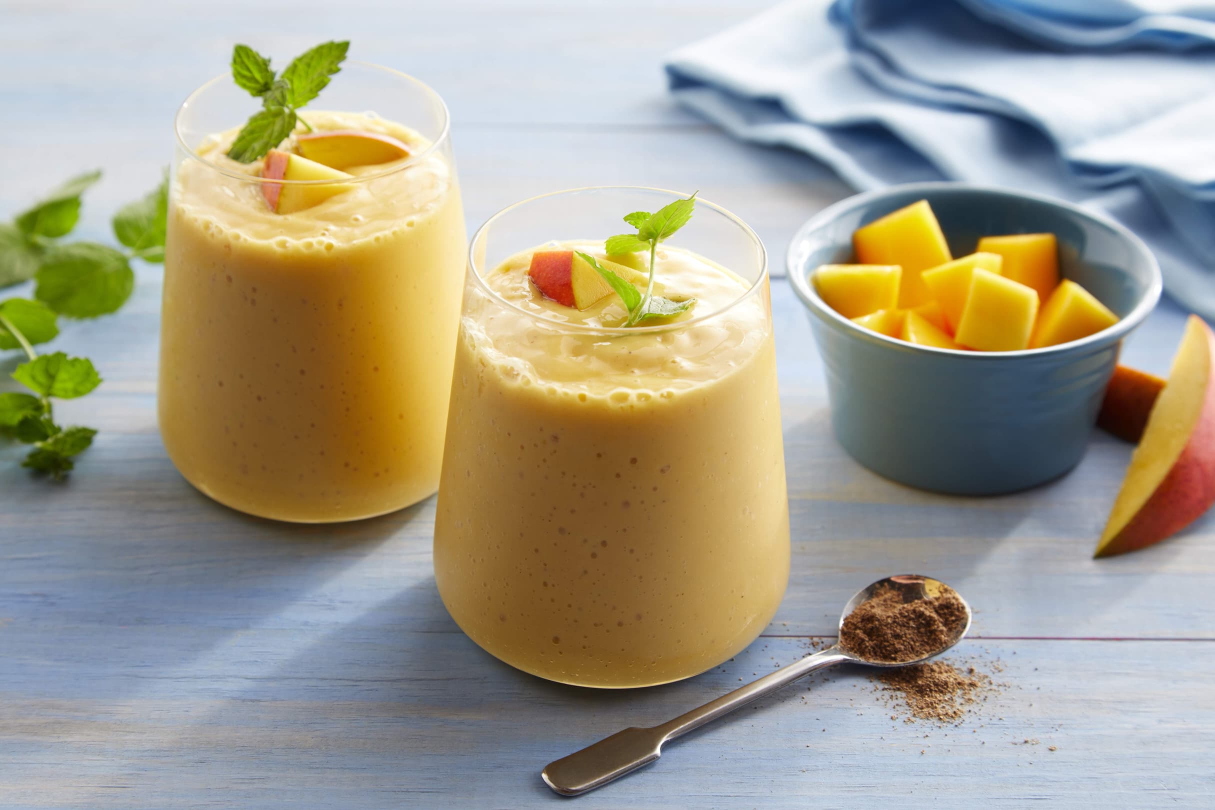Two glasses of mango smoothie topped with mint and mango slices, next to a bowl of diced mango and a spoon with ground spice, on a light blue table.