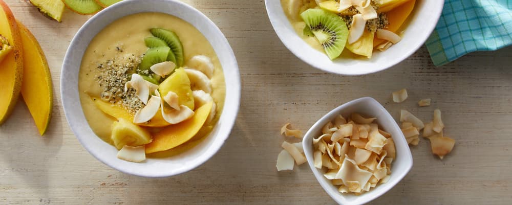 Two smoothie bowls topped with sliced mango, kiwi, banana, seeds, and toasted coconut flakes, next to a small bowl of coconut flakes and a blue cloth on a light wooden surface.