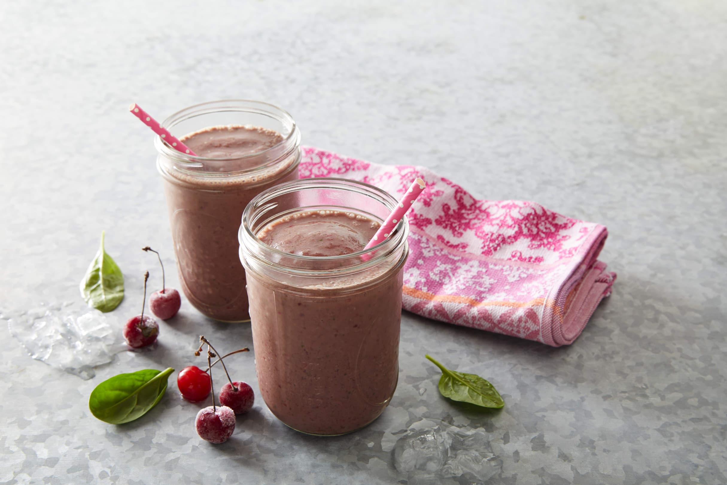 Two jars of pink smoothie with a straw, placed on a gray surface near spinach leaves, cherries, ice cubes, and a folded pink patterned cloth.