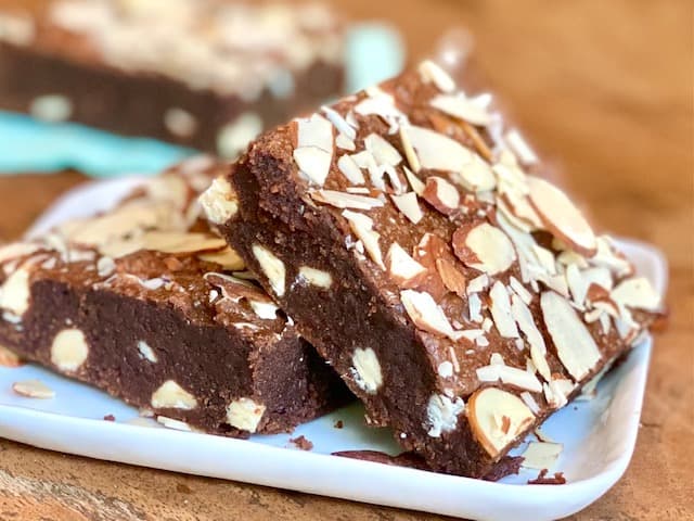 Two chocolate brownies topped with sliced almonds are stacked on a white plate, with more brownies visible in the blurred background.