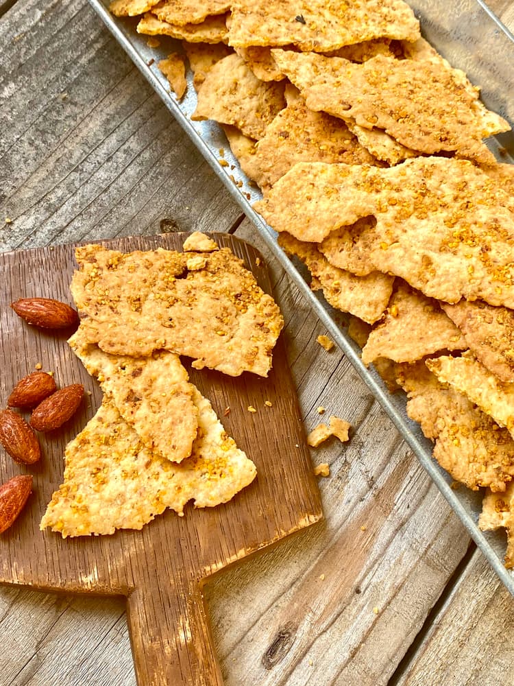 Crispy cracker pieces and roasted almonds are arranged on a wooden board and a metal tray, set on a rustic wooden table.