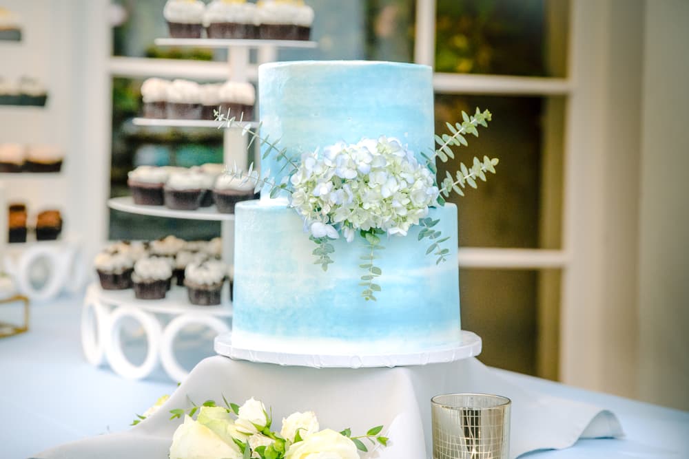 A two-tiered blue cake with white hydrangea flowers and eucalyptus leaves sits on a table, surrounded by cupcakes and white roses.