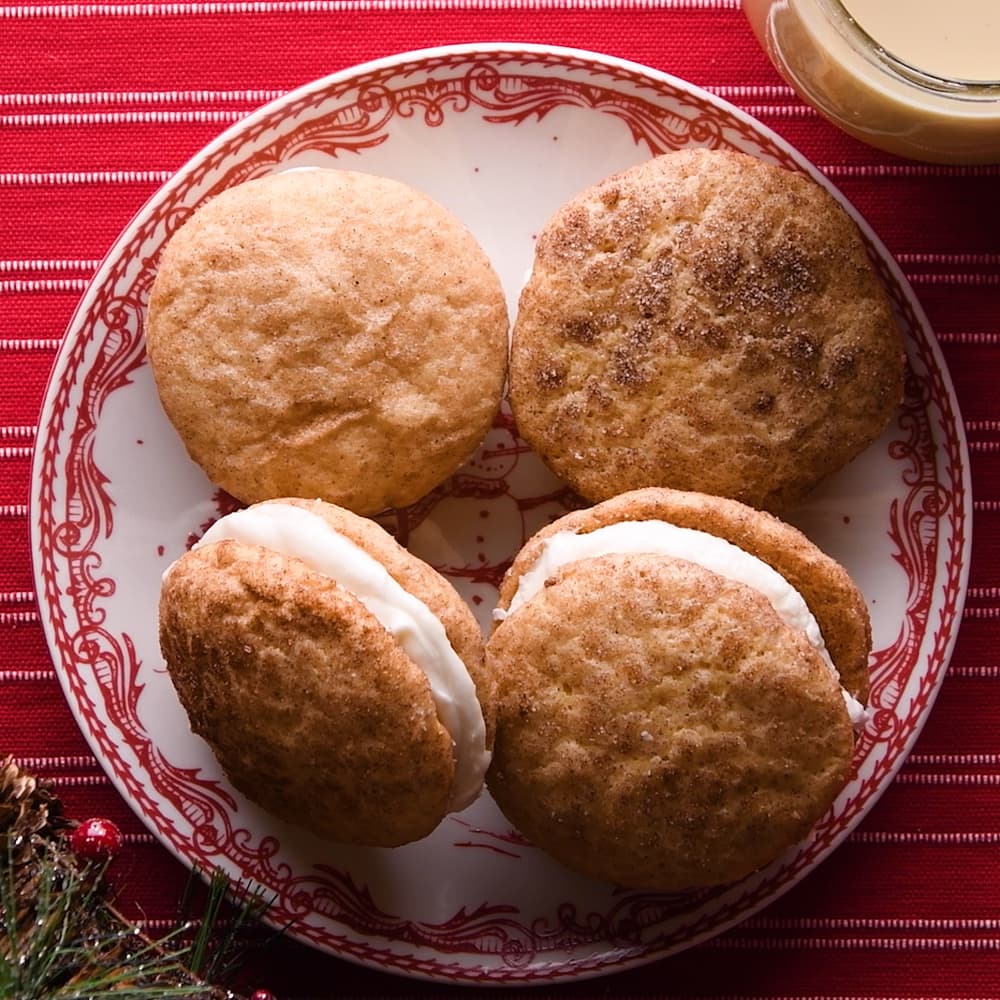 Four snickerdoodle whoopie pies with cream filling are arranged on a red and white plate, set on a red striped tablecloth with a festive holiday decoration visible in the corner.