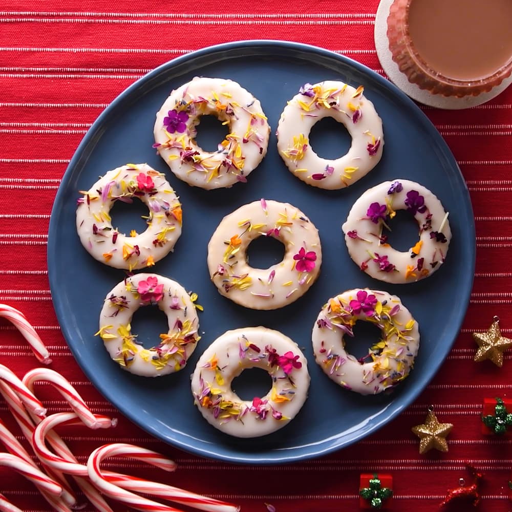 A blue plate with nine round, white-iced cookies topped with colorful flower petals, next to a cup of cocoa, candy canes, and gold star ornaments on a red striped tablecloth.