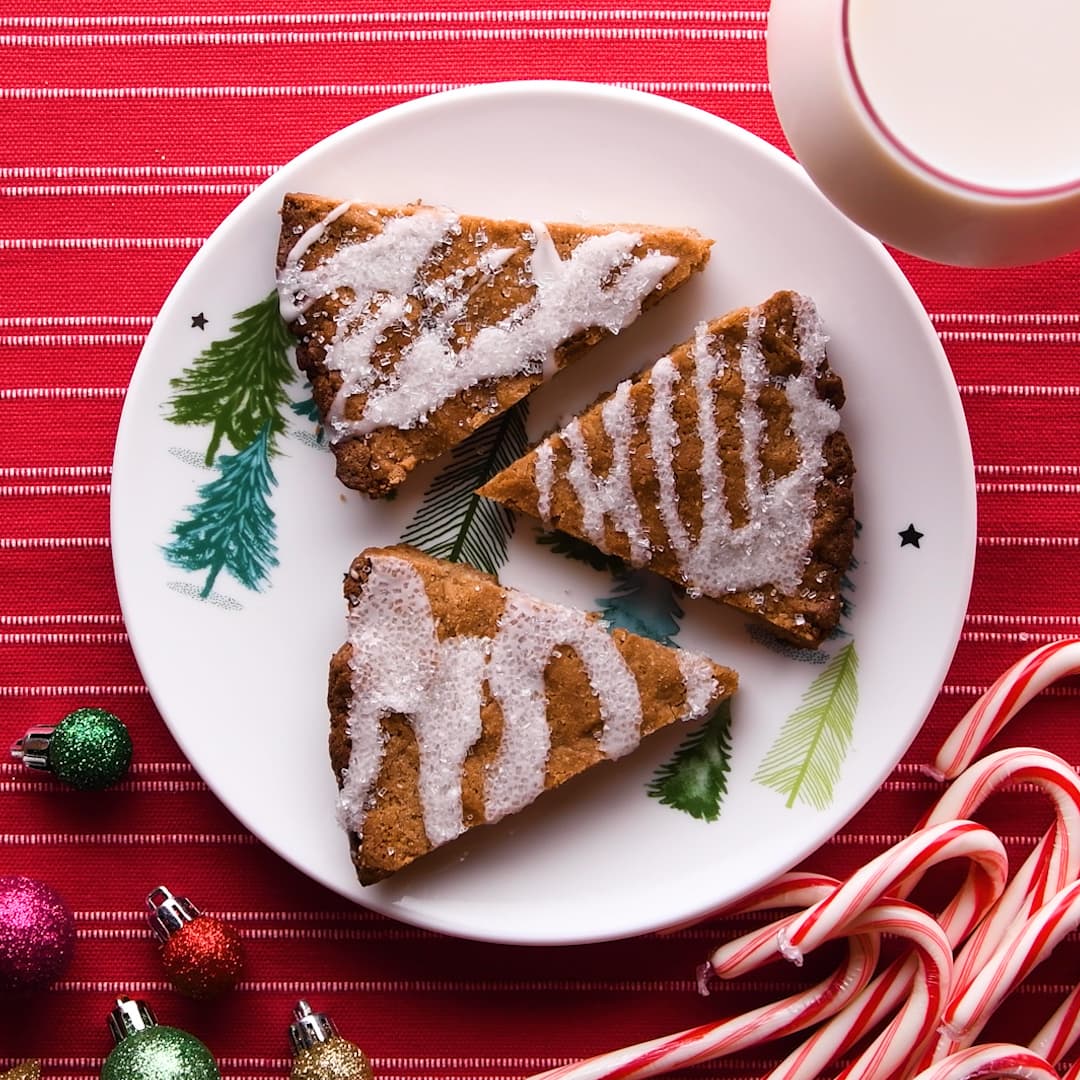 Three triangular pieces of iced cake on a holiday-themed plate, next to a glass of milk, candy canes, and small ornaments on a red striped tablecloth.