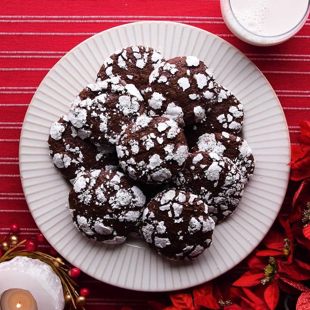 A white plate with chocolate crinkle cookies topped with powdered sugar, set on a red striped tablecloth, with a glass of milk and holiday decorations nearby.