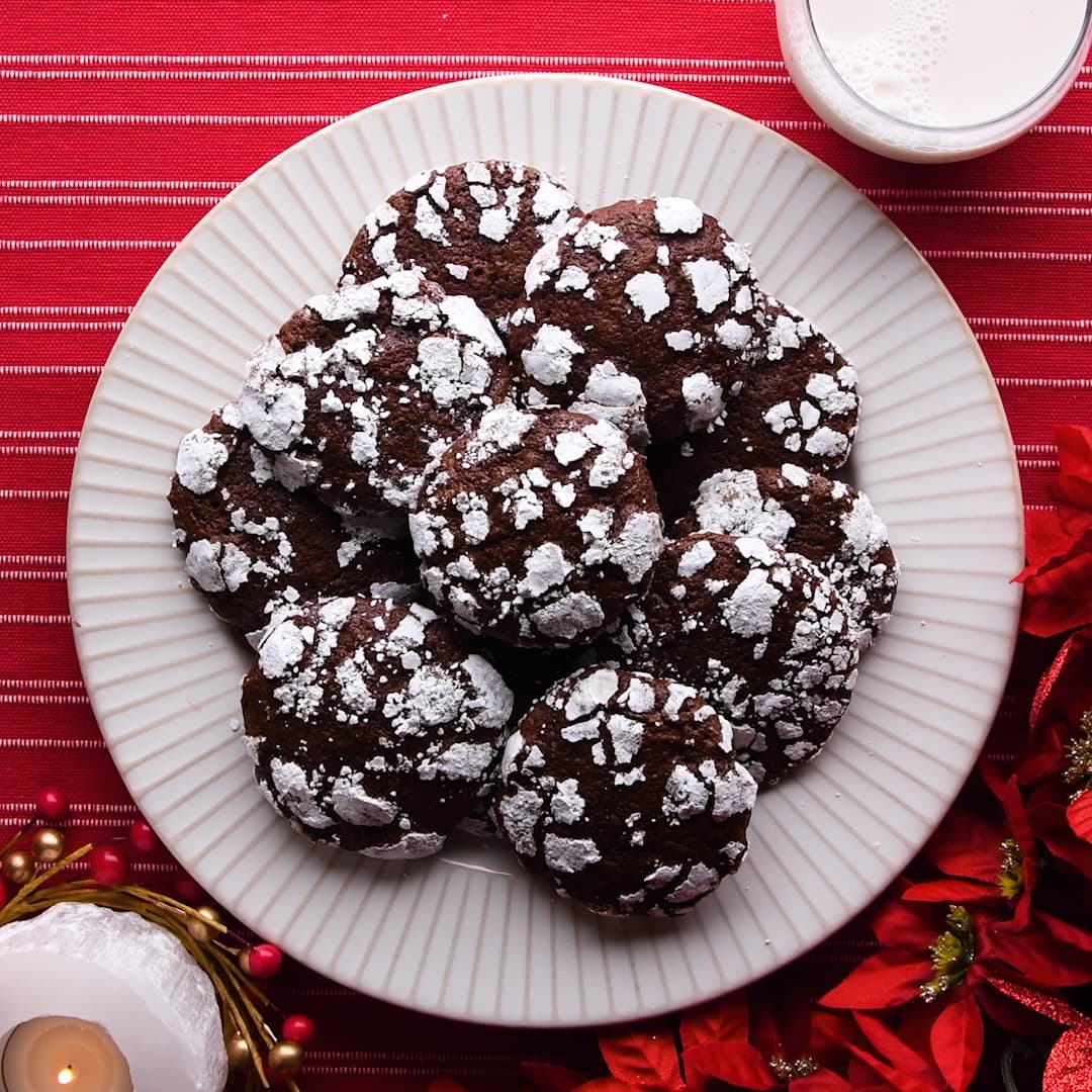 A white plate with chocolate crinkle cookies topped with powdered sugar, set on a red striped tablecloth, with a glass of milk and holiday decorations nearby.