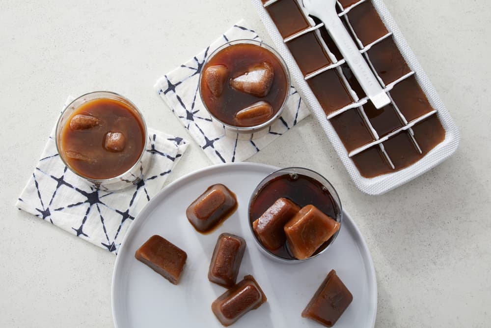 Three glasses with iced coffee and coffee ice cubes, a plate with extra ice cubes, and an ice cube tray filled with coffee on a light surface.