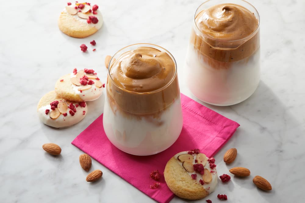 Two glasses of whipped coffee on milk sit on a pink napkin with almond cookies, sliced almonds, and dried berries on a marble surface.