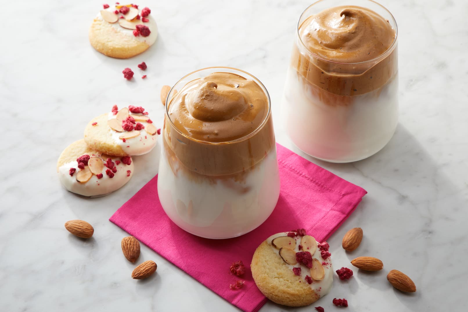 Two glasses of whipped coffee on milk sit on a pink napkin with almond cookies, sliced almonds, and dried berries on a marble surface.