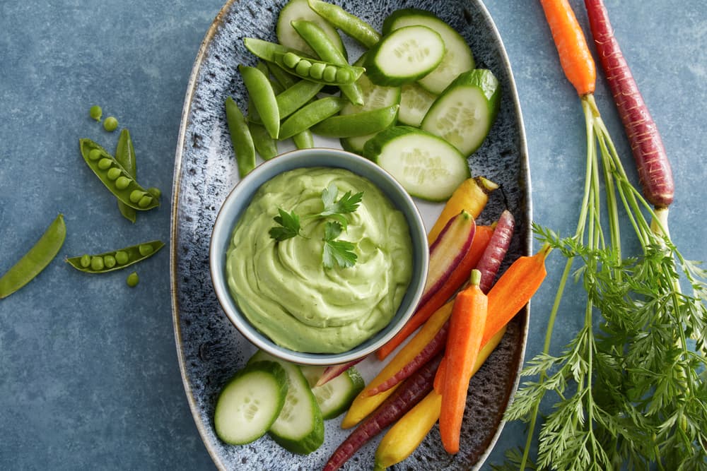 An oval plate with sliced cucumbers, snap peas, and colorful carrots arranged around a bowl of green dip, garnished with parsley.