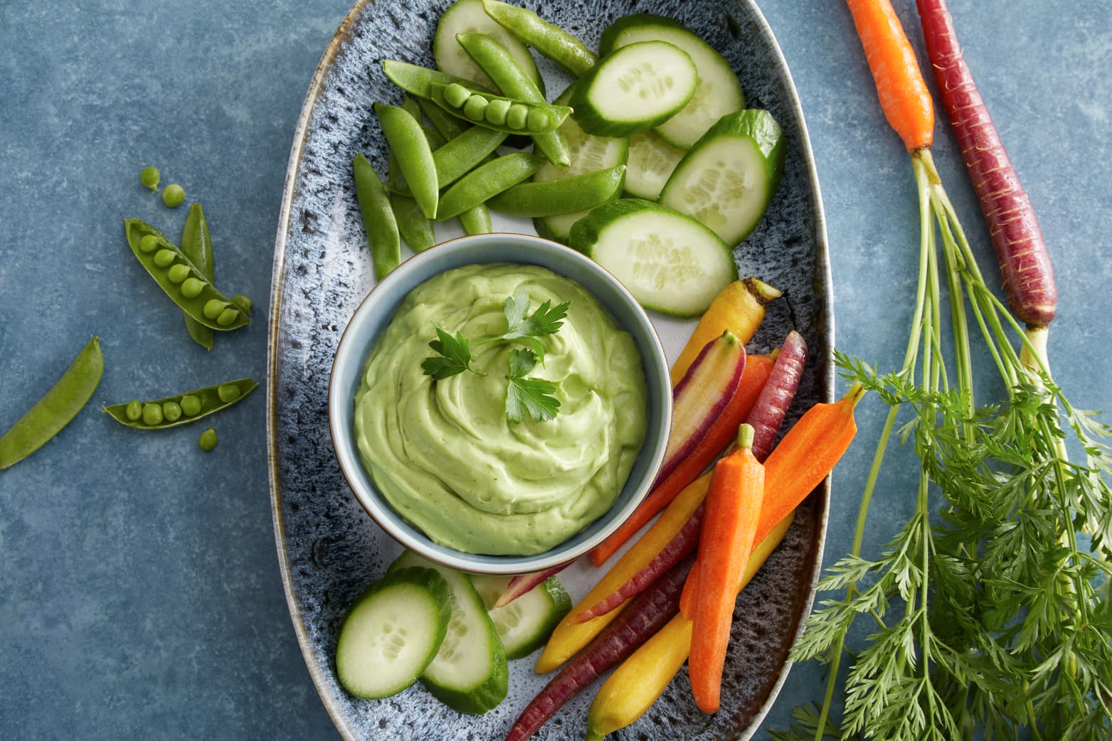 An oval plate with sliced cucumbers, snap peas, and colorful carrots arranged around a bowl of green dip, garnished with parsley.