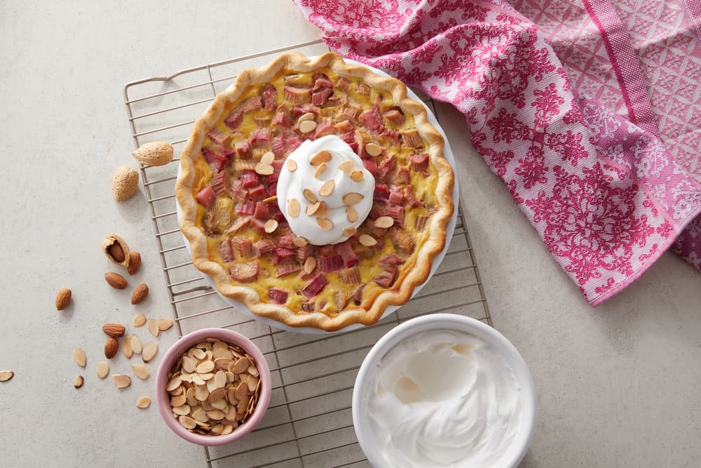 A baked pie with rhubarb and almonds topped with whipped cream sits on a cooling rack, next to a pink cloth, a bowl of sliced almonds, and a bowl of whipped cream.