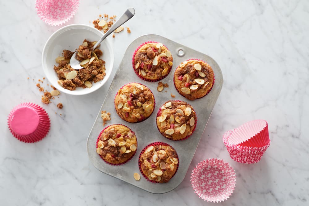 Six muffins topped with granola and sliced almonds in a tin, surrounded by pink paper liners and a bowl of granola on a marble surface.