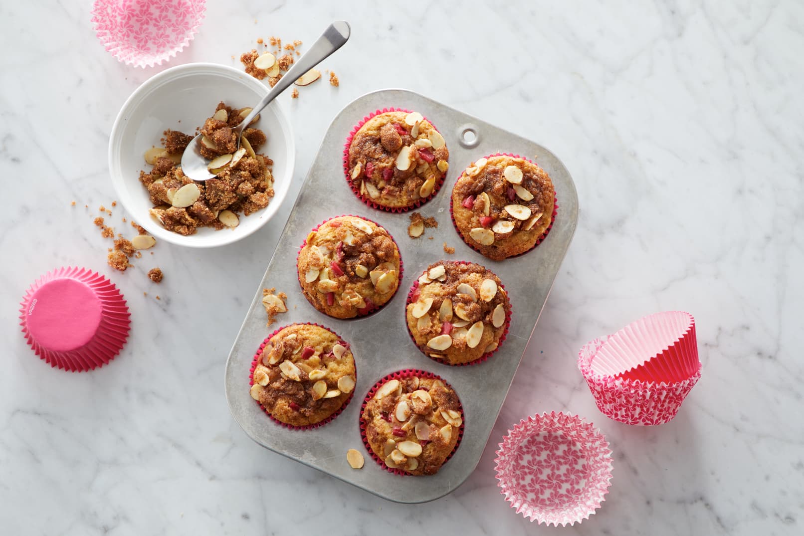 Six muffins topped with granola and sliced almonds in a tin, surrounded by pink paper liners and a bowl of granola on a marble surface.