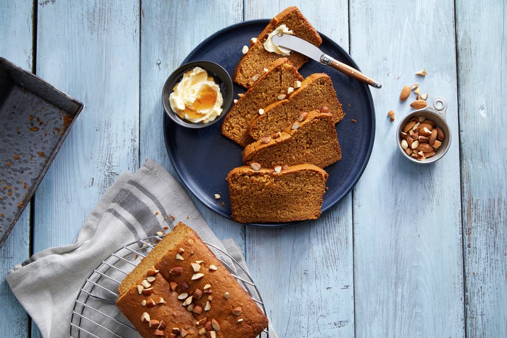 Sliced loaf cake on a dark plate with butter, a knife, and chopped nuts on a blue wooden table. A cooling rack and loaf pan are nearby.