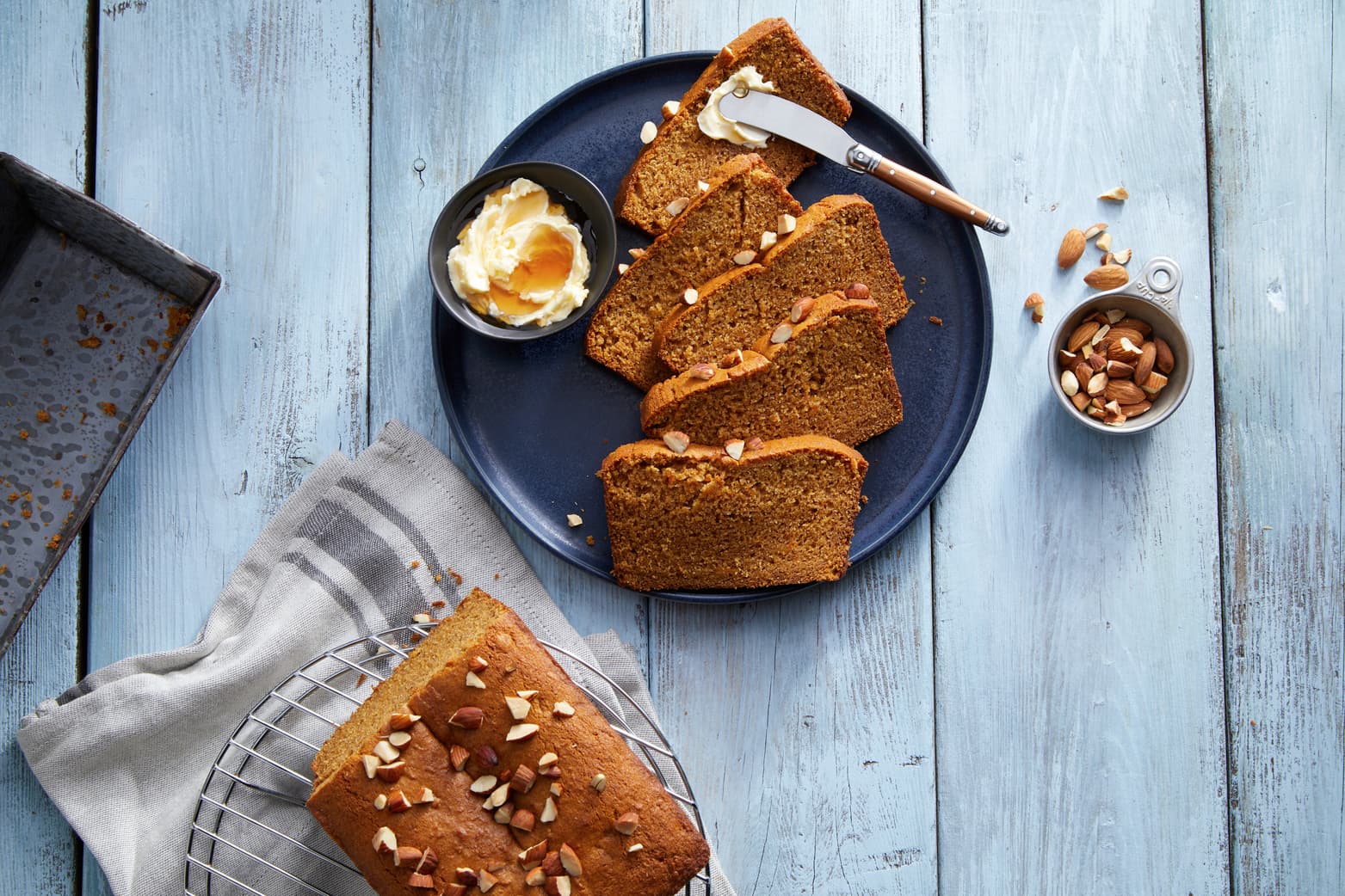 Sliced loaf cake on a dark plate with butter, a knife, and chopped nuts on a blue wooden table. A cooling rack and loaf pan are nearby.