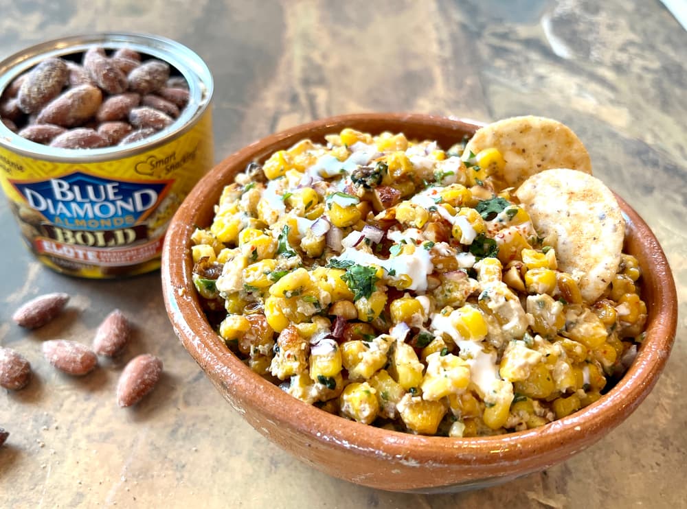 A bowl of seasoned corn salad with herbs and sauce, garnished with two crackers, sits next to a can of Blue Diamond Bold almonds on a brown tabletop.