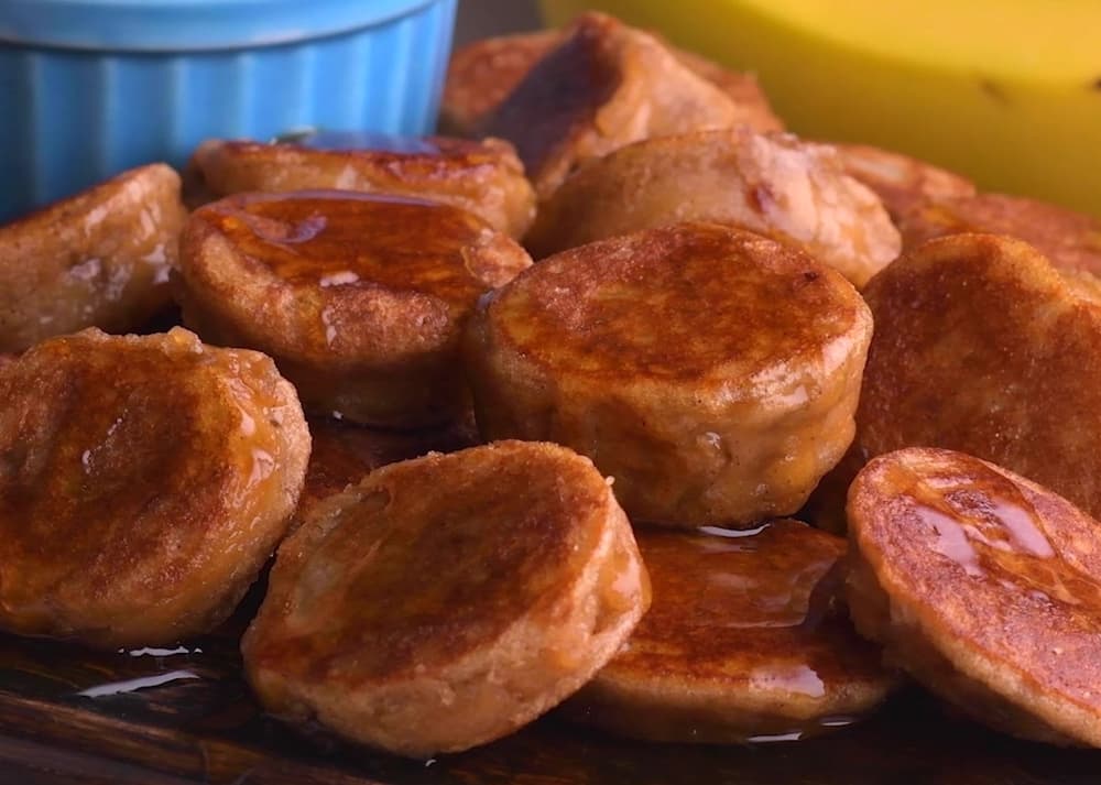 A pile of golden-brown banana pancakes drizzled with syrup, placed on a wooden surface with part of a blue bowl and a banana visible in the background.