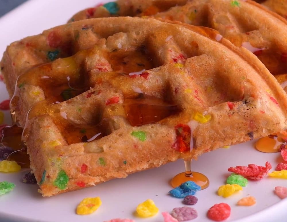 A close-up of two colorful waffles with syrup drizzled on top, served on a white plate with pieces of fruity cereal scattered around.