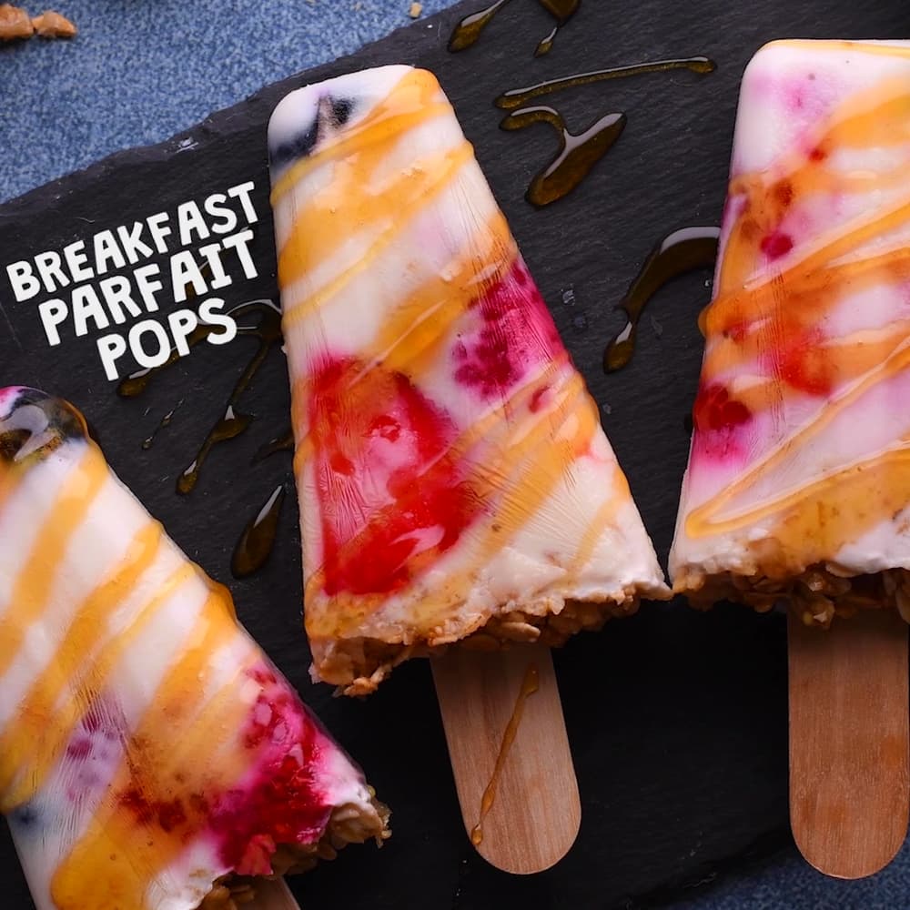 Three yogurt parfait popsicles with fruit and granola are arranged on a dark surface, drizzled with honey. Text on the image reads Breakfast Parfait Pops..