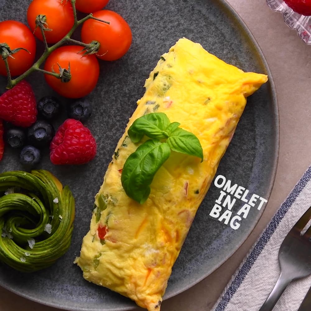 A rectangular omelet garnished with basil is served with tomatoes on the vine, raspberries, blueberries, and a sliced avocado on a dark plate. Text reads OMELET IN A BAG..