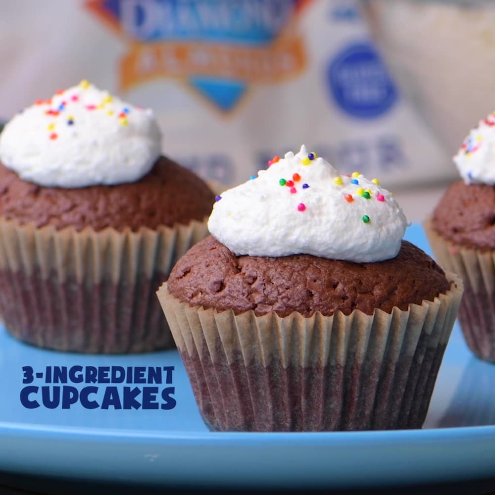 Three chocolate cupcakes with white whipped topping and colorful sprinkles sit on a blue plate. The text 3-INGREDIENT CUPCAKES appears in the corner.