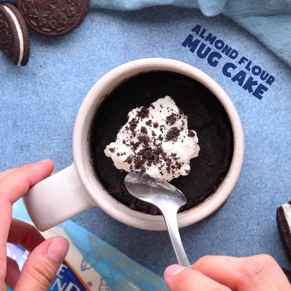 A hand holds a spoon over a mug filled with chocolate almond flour mug cake, topped with whipped cream and cookie crumbs.