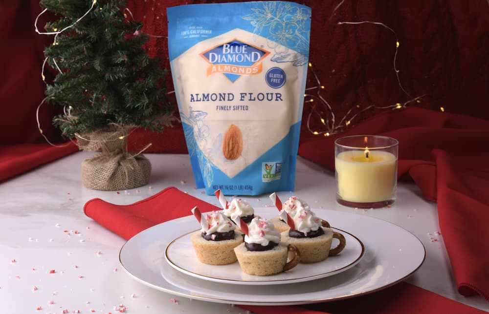 A plate of mini dessert cups with whipped cream and crushed peppermint, a bag of almond flour, a lit candle, and a small Christmas tree on a table with red napkins.