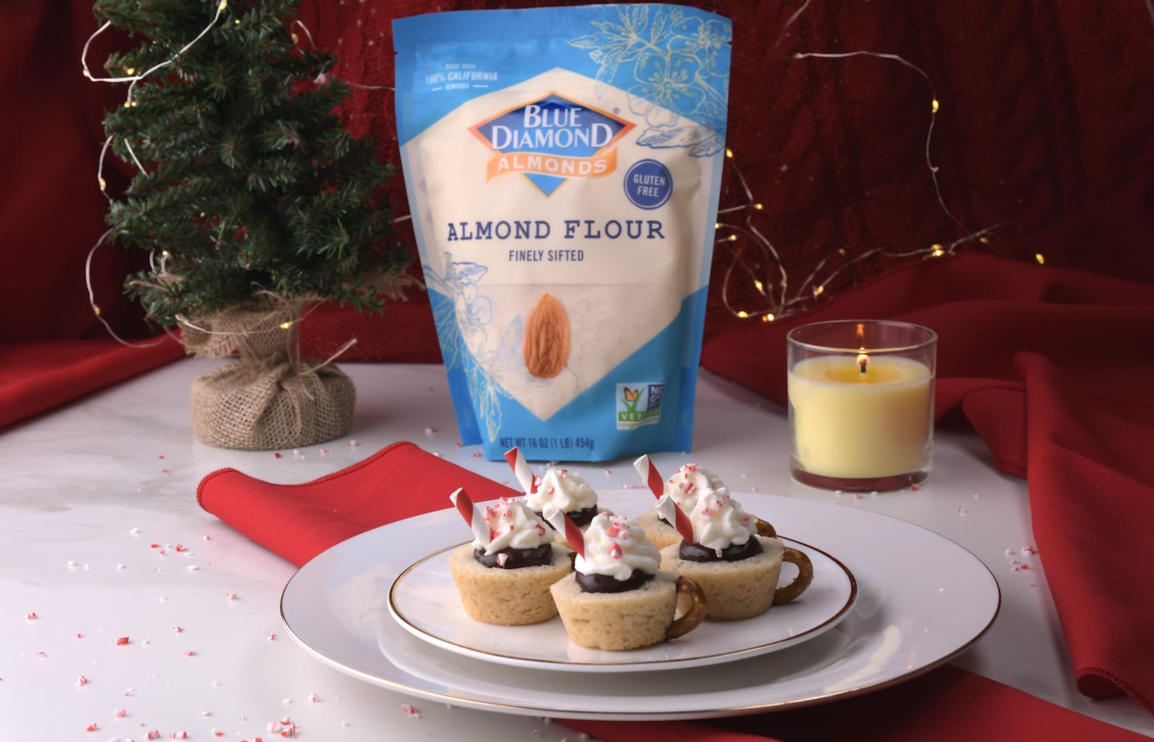 A plate of mini dessert cups with whipped cream and crushed peppermint, a bag of almond flour, a lit candle, and a small Christmas tree on a table with red napkins.