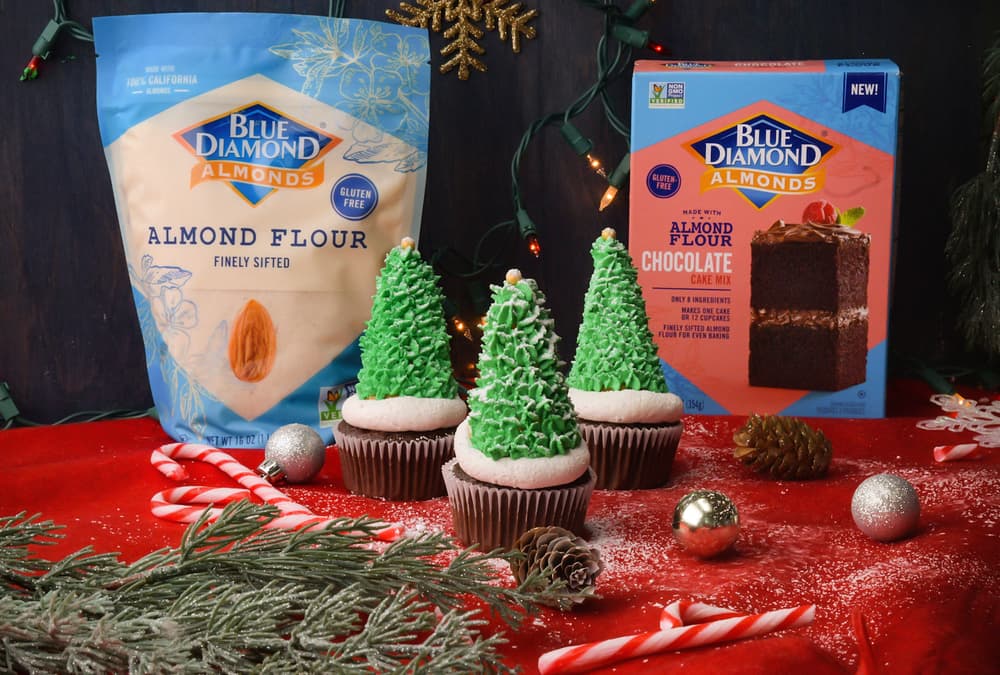 Three cupcakes decorated as Christmas trees on a red cloth, with Blue Diamond Almond Flour products, candy canes, pine branches, and ornaments in the background.