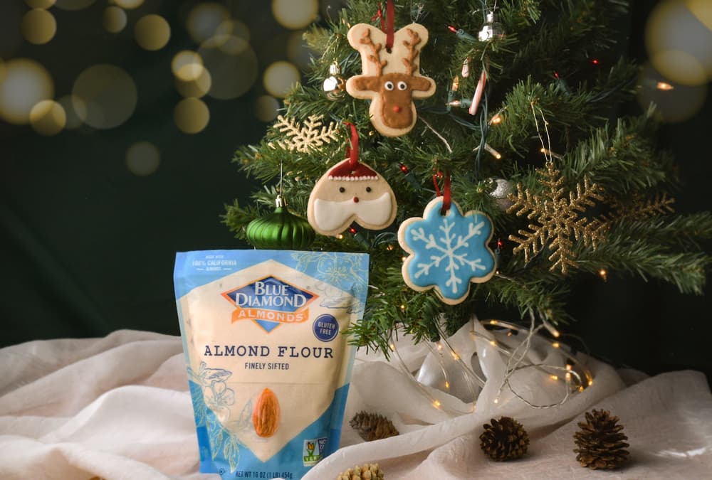 A bag of Blue Diamond almond flour sits in front of a decorated Christmas tree with cookie ornaments and pinecones on a white cloth.
