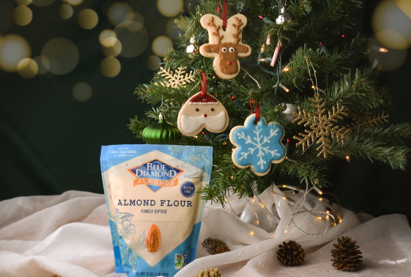 A bag of Blue Diamond almond flour sits in front of a decorated Christmas tree with cookie ornaments and pinecones on a white cloth.