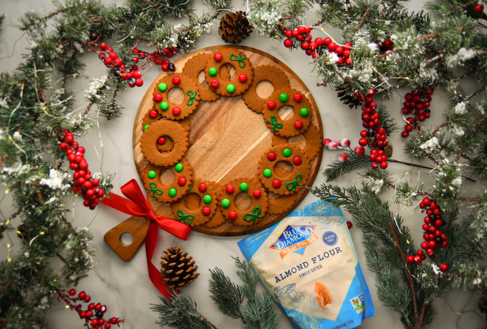A round wooden board with decorated holiday cookies, a red ribbon, almond flour, pinecones, artificial snow, and red berries arranged in a festive display.