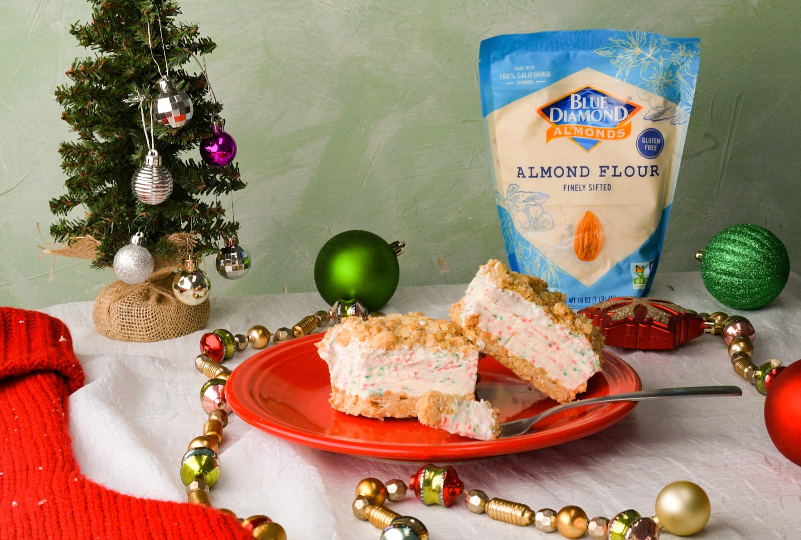 A red plate with two slices of dessert sits beside a fork, Christmas decorations, and a bag of Blue Diamond Almond Flour on a festive table.